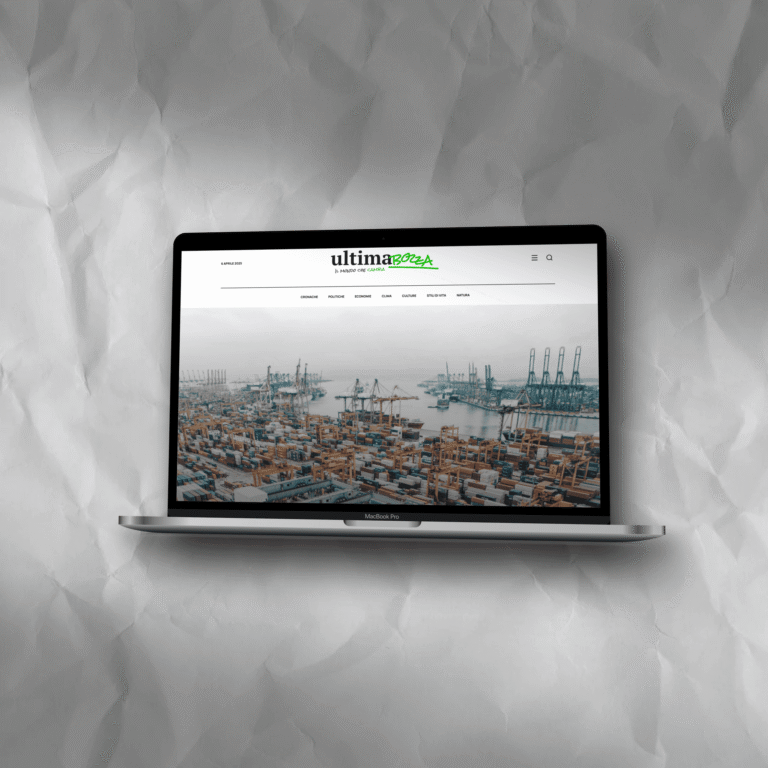 Un computer portatile MacBook Pro è mostrato su uno sfondo grigio chiaro e stropicciato. Lo schermo del laptop visualizza la homepage di un sito web chiamato "ultimabozza". La homepage presenta un'immagine di sfondo di un porto industriale trafficato, pieno di container colorati, navi e gru portuali. La barra di navigazione superiore del sito web è bianca e contiene il logo "ultima[bozza]" in nero e verde a sinistra, diverse voci di menu e un'icona di ricerca e un'icona a tre linee (menu hamburger) a destra.